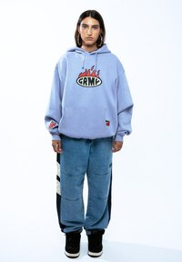 Sudadera de color púrpura claro con un gráfico de llamas, bolsillo frontal y logo. Combinada con pantalones de mezclilla holgados de color azul y zapatillas altas negras.