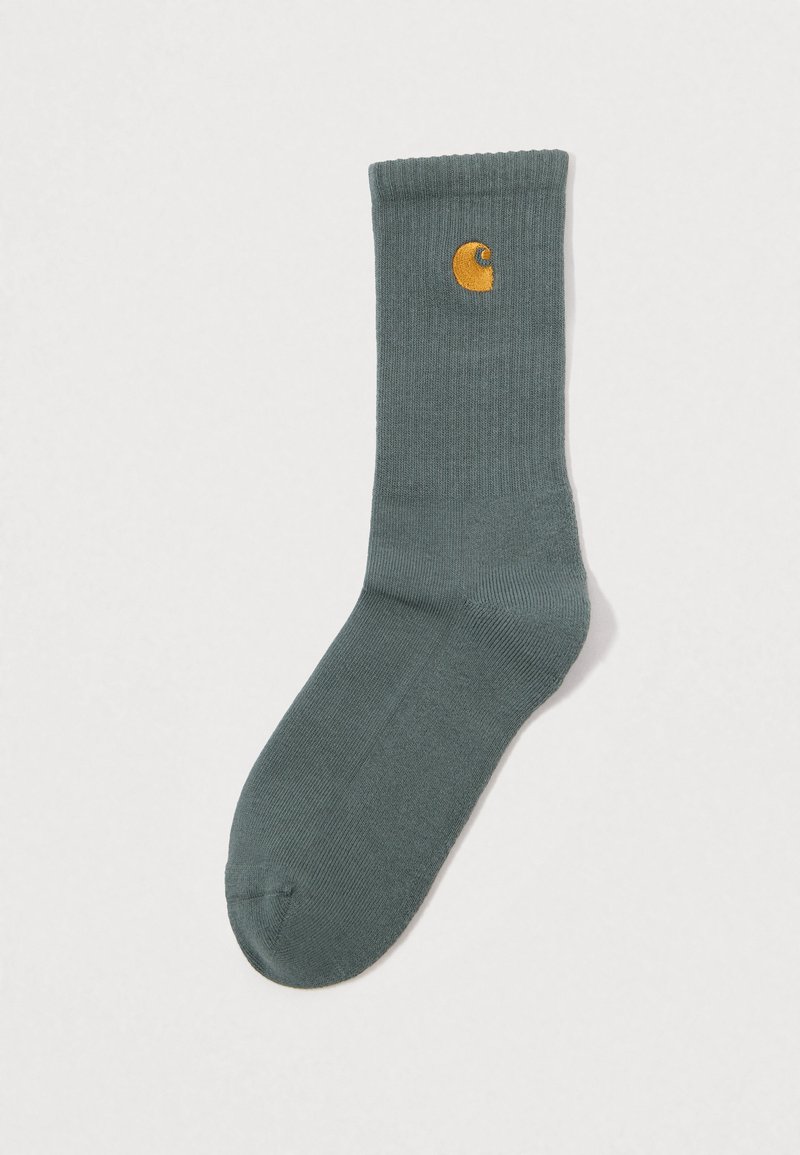 Calcetín gris de crew, con puño acanalado y pequeño logo de Carhartt bordado en amarillo cerca de la parte superior, tendido en un fondo blanco.