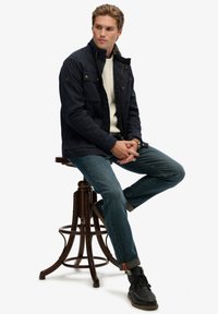 Marineblauwe jacket met rits en drukknoopsluiting, vier voorzakken, gedragen over een crèmekleurige trui, gecombineerd met blauwe jeans en zwarte laarzen.