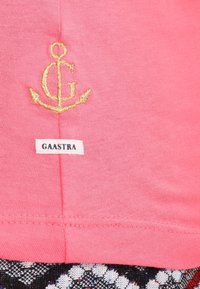 Korallenrosa Baumwoll-T-Shirt mit goldfarbener Anker-Logo-Stickerei und einem Etikett mit "GAASTRA" in Schwarz auf weißem Hintergrund.