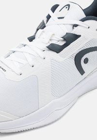 Scarpa sportiva bianca con tomaia sintetica testurizzata, dettagli scuri e sistema di allacciatura tradizionale. Presenta una suola ammortizzata e aree in mesh traspirante.