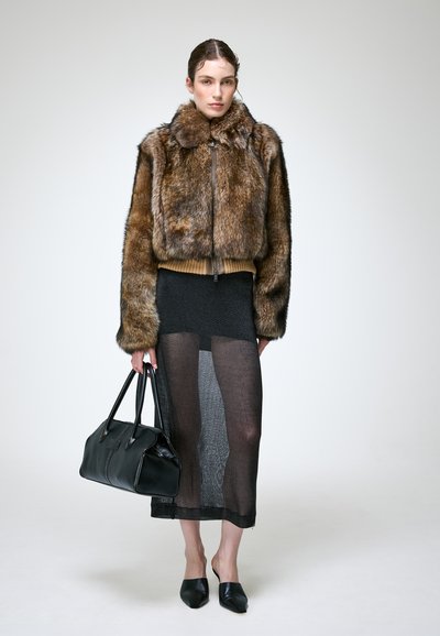 Femme portant une veste en fourrure marron, une jupe noire transparente, des chaussures noires pointues, portant un sac à main noir sur un fond clair uni.