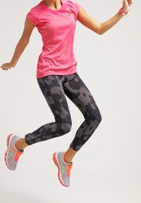 Camiseta atlética ligera rosa, leggins negros con estampado y zapatillas de correr grises con detalles en naranja. Modelo en pleno salto.