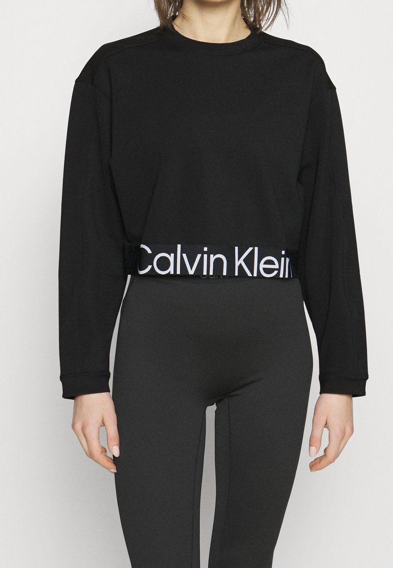 Personne portant un crop top noir à manches longues avec bande blanche logo Calvin Klein et des leggings noirs taille haute sur fond blanc.