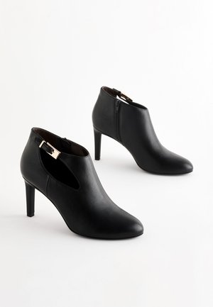 Schwarze Leder-Stiefeletten mit spitzen Zehen, schlanken hohen Absätzen, seitlichen Ausschnitten und kleinen metallischen Schnallen, mit einem Reißverschluss an der Rückseite.