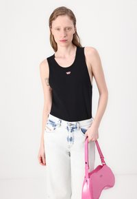 Crna majica bez rukava s malim crvenim logom, u kombinaciji s svijetloplavim trapericama visokog struka sa suptilnim tie-dye uzorkom i sjajnom ružičastom torbicom.