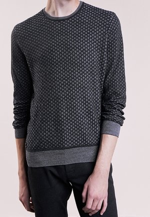 Person iført en langærmet, mørkegrå mønstret sweater med ribstrikkede manchetter og talje, kombineret med sorte bukser.