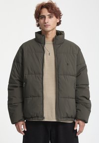 Chaqueta de plumón en verde oliva oscuro, con un alto cuello, bolsillos laterales con cremallera y textura acolchada. Llevada sobre una camisa beige clara.