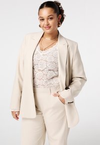 Jonge vrouw met krullend haar in een beige pak en een wit kant top, glimlachend met een hand in haar zak tegen een effen achtergrond.