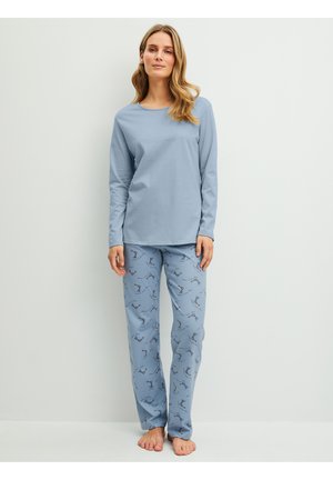 Woman standing barefoot wearing light blue long-sleeve top and patterned pajama pants with bird illustrations against a plain light background.