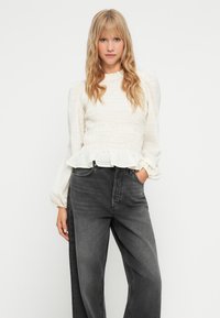 Blusa bianca arricciata con maniche a palloncino e orlo a volant, abbinata a jeans a gamba larga grigio scuro a vita alta. Tessuto morbido, vestibilità rilassata.