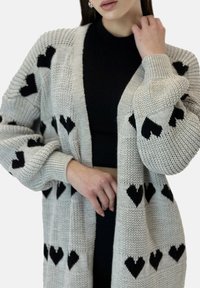 Szary dzianinowy kardigan z wyraźnymi czarnymi wzorami w kształcie serc, długimi rękawami i prążkowanymi mankietami. Otwarty przód z fakturowanym wykończeniem.