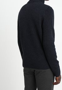 Mörkblå stickad tröja med hög krage, texturerad tyg, ribbat nederkant och ärmslut, bärs med randiga byxor.