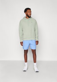 Lys grønn hettegenser med snøring og logo, kombinert med blå treningsshorts. Hvite joggesko med grå detaljer, stående mot en enkel bakgrunn.