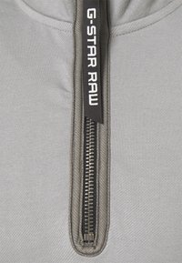 Gros plan d'une fermeture éclair grise avec une tirette noire comportant un texte blanc "G-STAR RAW".