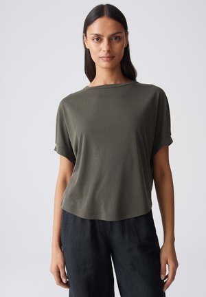 Frau mit langen dunklen Haaren, die ein locker sitzendes olivgrünes Kurzarmshirt und schwarze Hosen trägt, steht vor hellem grauem Hintergrund.