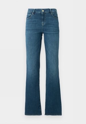 Donna con occhiali da sole neri, top bianco senza maniche, jeans blu con cintura nera e ballerine nere a punta, in piedi con una mano in tasca.