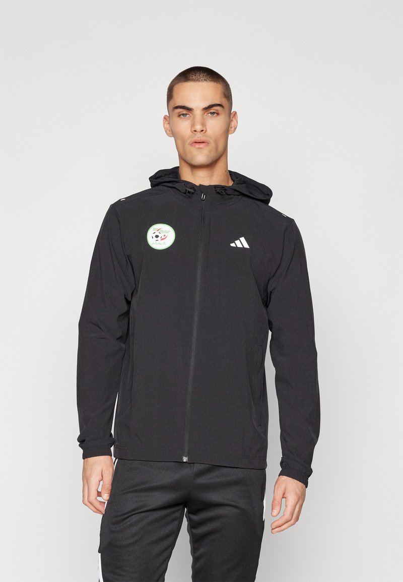Crna jakna sa patentnim zatvaračem i kapuljačom, sa zelenom kružnom oznakom na prsima i bijelim detaljima logotipa Adidas. Napravljena od laganog materijala.
