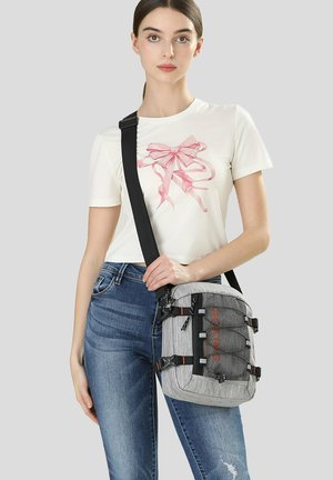 Vrouw draagt een wit t-shirt met een roze strikafbeelding, een blauwe spijkerbroek en heeft een grijze crossbodytas met zwarte band en een voorvak van gaas bij zich.