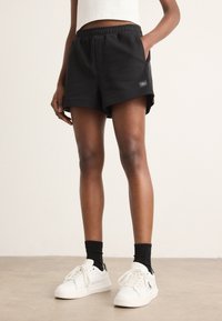 Person iført sorte shorts med elastisk talje, sorte sokker og hvide sneakers står på en glat lysebrun gulv mod en ensfarvet væg.