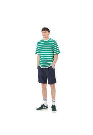 Camiseta de rayas verdes y blancas de corte holgado, combinada con pantalones cortos azul marino. Se lleva con zapatillas verde oscuro y calcetines tobilleros con rayas negras.