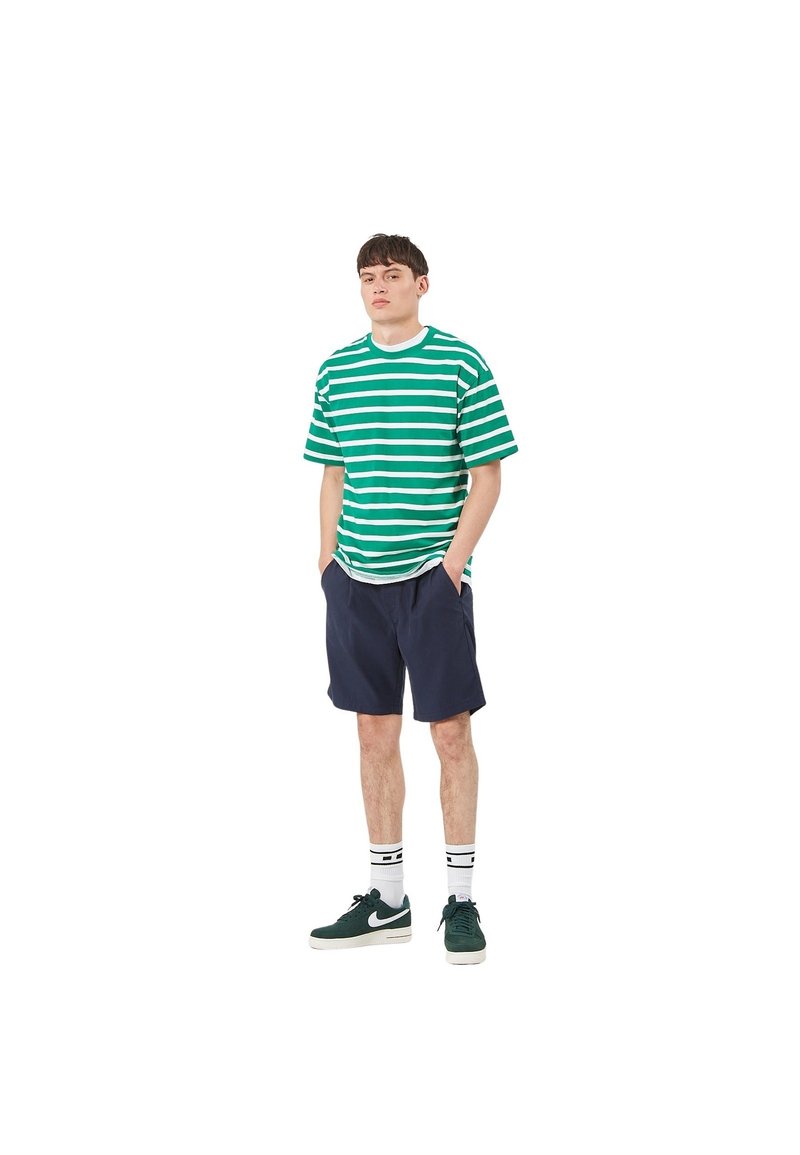 Maglietta a righe verdi e bianche con vestibilità ampia, abbinata a pantaloncini blu navy. Indossata con scarpe da ginnastica verde scuro e calzini alla caviglia con strisce nere.