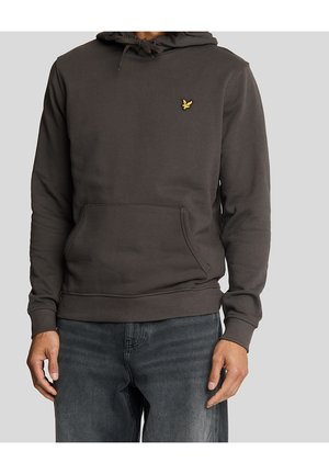 Mand iført mørkegrå hættetrøje med en lille gul fugle-logo på venstre bryst, kombineret med mørk vaskede jeans.