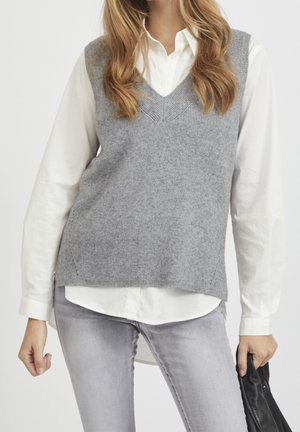 Femme portant un pull sans manches en maille grise par-dessus une chemise blanche boutonnée et un jean gris clair, tenant un vêtement noir dans la main droite.