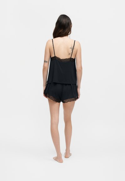 Γυναίκα που στέκεται ξυπόλητη, φοράει μαύρο καμιζόλε με δαντέλα και σορτς πιτζάμες, από πίσω σε απλό φόντο.