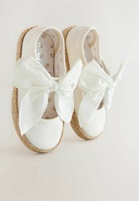 Scarpe per bambini bianche con grandi fiocchi di stoffa, suole in giuta intrecciata e fodera interna a motivi floreali su uno sfondo semplice.