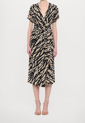 Femme portant une robe midi portefeuille beige et noire à imprimé abstrait avec manches courtes et talons nus à bride cheville, posant sur fond blanc.