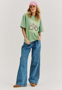 Woman wearing a pink cap, green oversized t-shirt with bicycle print, wide-leg blue jeans, and brown shoes, standing against a plain background.