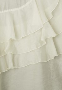 Tissu couleur crème avec des volants superposés. Texture translucide et lisse avec une finition délicate et un léger éclat. Les bords présentent de fins détails de couture.