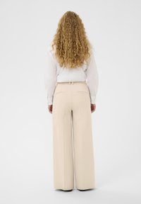 Pantalon large beige avec une coupe ajustée, présentant une coupe droite, une texture subtile et deux poches arrière. Accompagné d'un chemisier blanc.