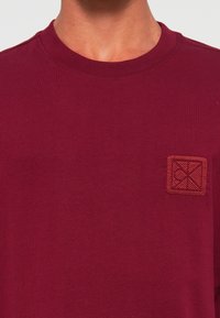 Hombre con una camiseta de cuello redondo burdeos con un pequeño emblema cuadrado bordado en el lado izquierdo del pecho.