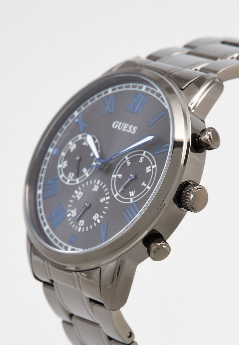 Męski zegarek Guess z srebrną metalową bransoletą, czarną tarczą, niebieskimi cyframi rzymskimi oraz trzema subtarczami pokazującymi datę, dzień tygodnia i czas w formacie 24-godzinnym.