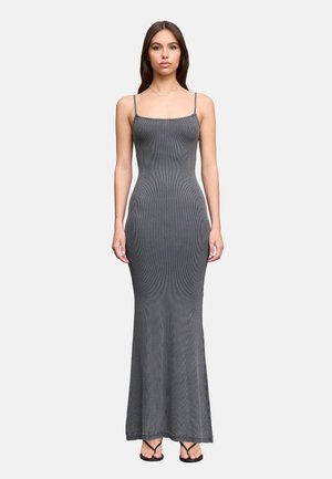 Donna in piedi che indossa un vestito grigio a coste aderente, senza maniche, lungo fino alla caviglia con sottili bretelle e sandali neri, rivolta in avanti su sfondo semplice.