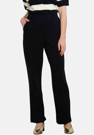 Mujer con pantalones negros de pierna recta, blusa a rayas negras y blancas, y zapatos dorados de tacón bajo, con una mano en el bolsillo.