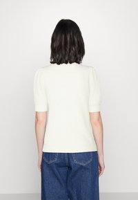 Trumpas rankovėmis, kreminės spalvos megztinis su tekstūruotu raštu ir raukšlėmis apkirptomis detalėmis, derinamas su aukštu liemeniu mėlynomis džinsų kelnėmis.