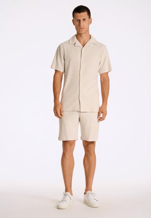 Mann steht vor weißem Hintergrund, trägt beige kurzärmeliges Hemd mit Knöpfen und passende Shorts sowie weiße Turnschuhe.