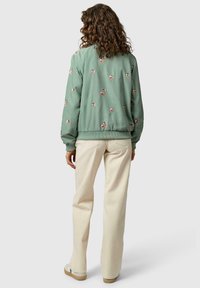 Kobieta o kręconych włosach nosi jasną zieloną kurtkę bomber z kwiatowym wzorem, kremowe spodnie i białe trampki, stoi tyłem na jednolitym tle.