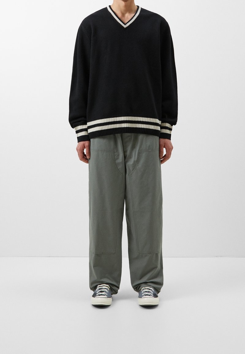Czarny sweter dzianinowy z dekoltem w kształcie V i białymi paskami na mankietach, zestawiony z luźnymi, jasnoszarymi spodniami. Teksturowana tkanina i swobodny krój.