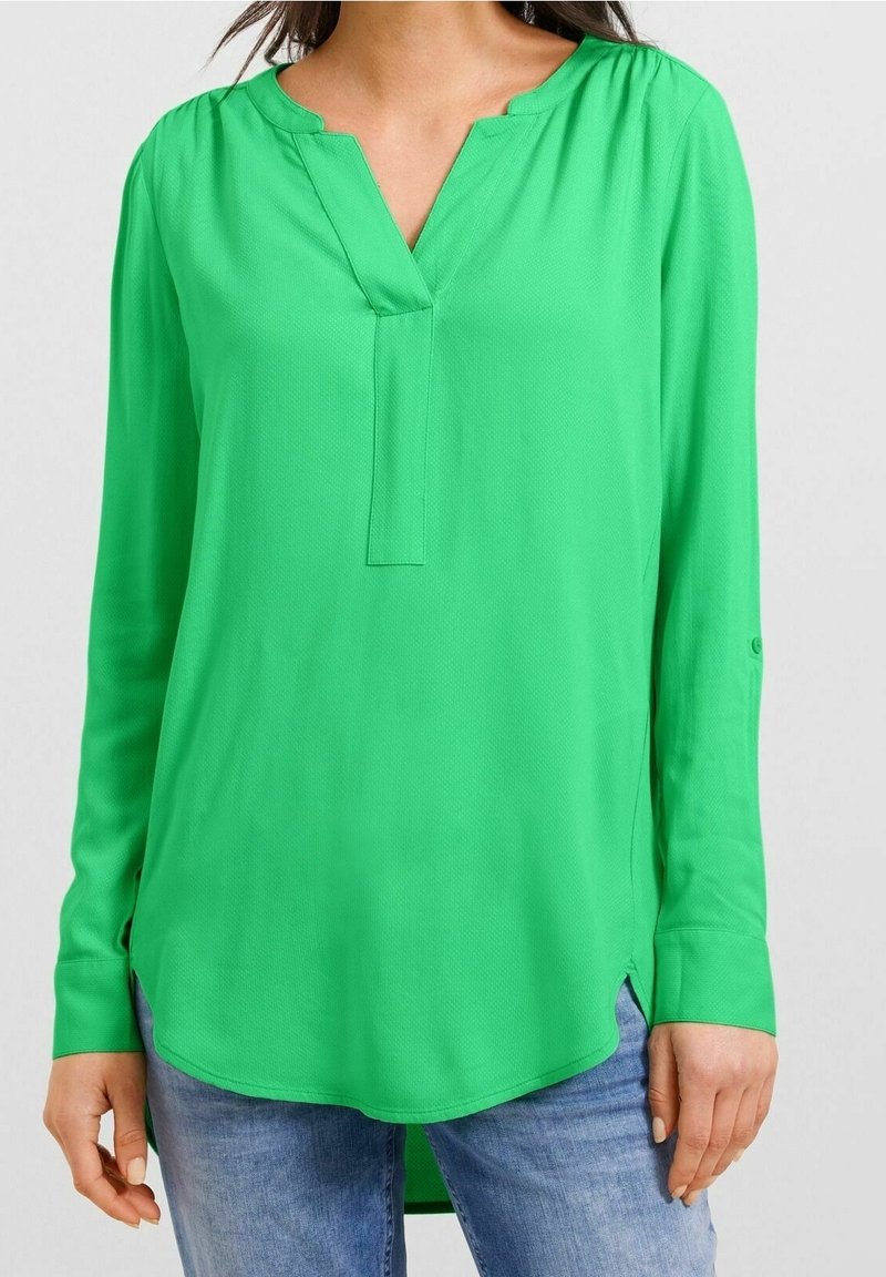 Femme portant un chemisier vert vif à manches longues avec un col en V et un jean bleu clair sur un fond neutre.