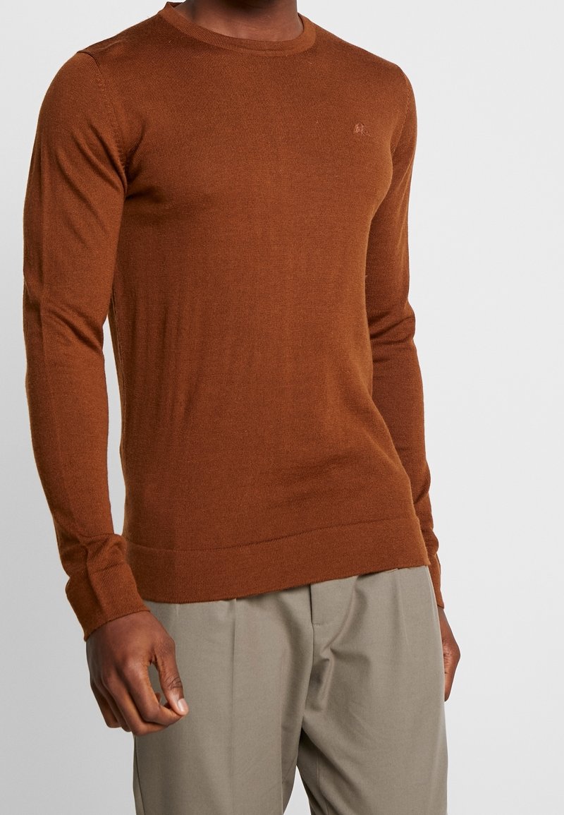 Homme portant un pull en maille manches longues couleur rouille associé à un pantalon beige clair sur un fond uni.
