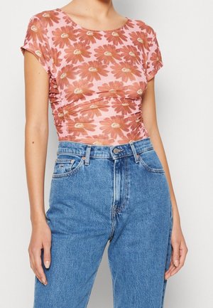 Femme portant un haut rose moulant avec de grandes fleurs orange associé à un jean denim bleu taille haute sur fond uni.