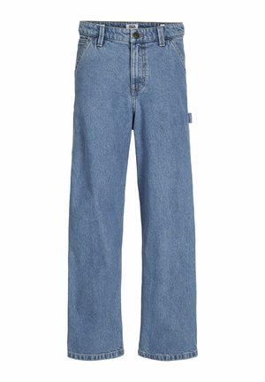 Personne portant un jean bleu ample, des baskets blanches et un t-shirt blanc, les mains dans les poches du jean, debout devant un fond uni.