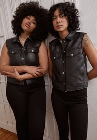 Coletes de couro preto com grandes bolsos frontais, botões de pressão e design sem mangas com arolhamento, usados sobre calças pretas ajustadas.