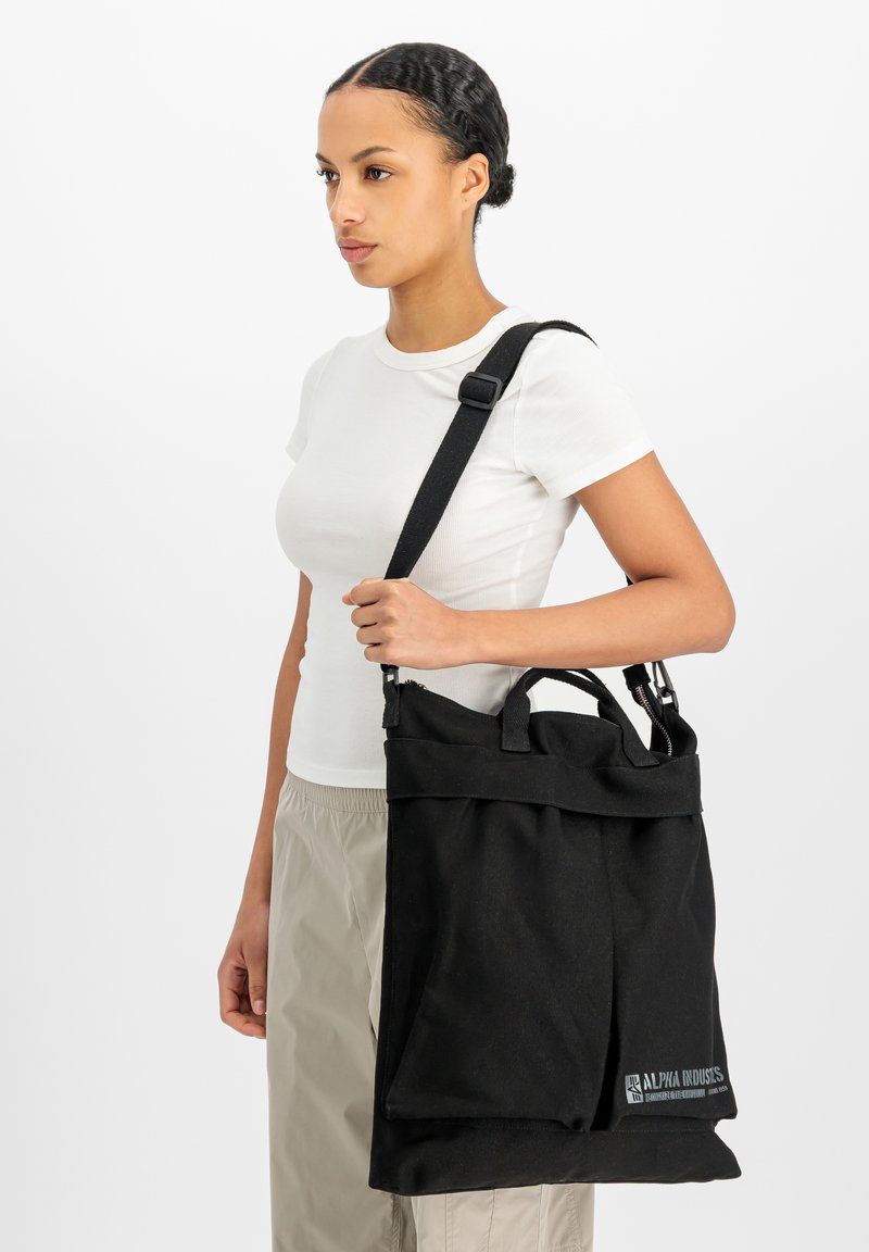 Femme portant une chemise blanche et un pantalon beige porte un grand sac cabas noir Alpha Industries avec une bandoulière.