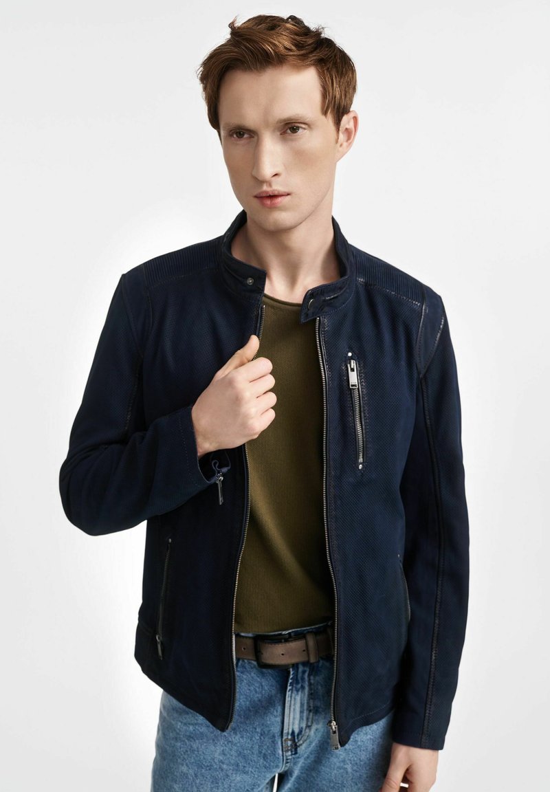 Granatowa zamszowa kurtka z wysokim kołnierzem, zamkiem błyskawicznym z przodu i kieszeniami. Nosi się ją na zieloną koszulę, w zestawieniu z jasnoniebieskimi dżinsami. Gładka faktura.