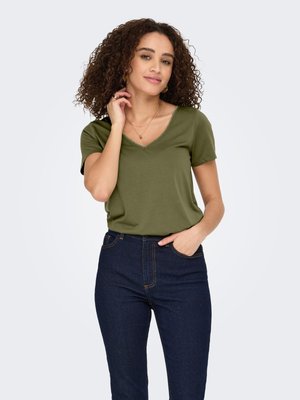 Femme aux cheveux bouclés portant un t-shirt vert olive à col en V à manches courtes et un jean bleu foncé, posant avec une main près du visage.